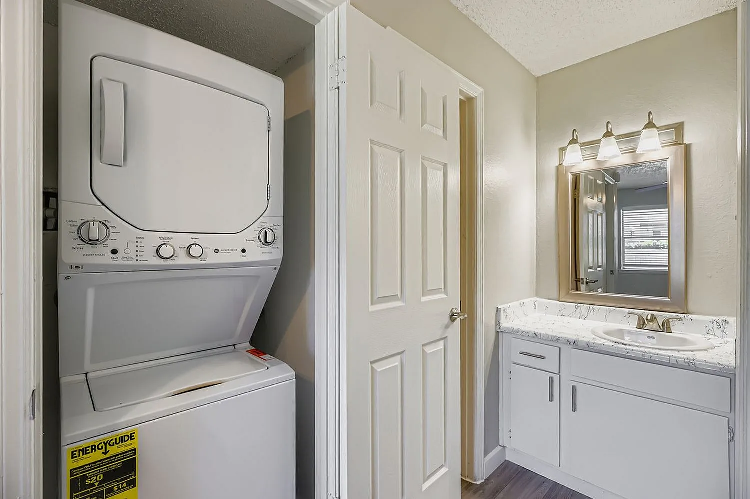 Laundry alcove with stacked washer-dryer unit, bathroom with white vanity, marble countertops, mirror with brass fixtures