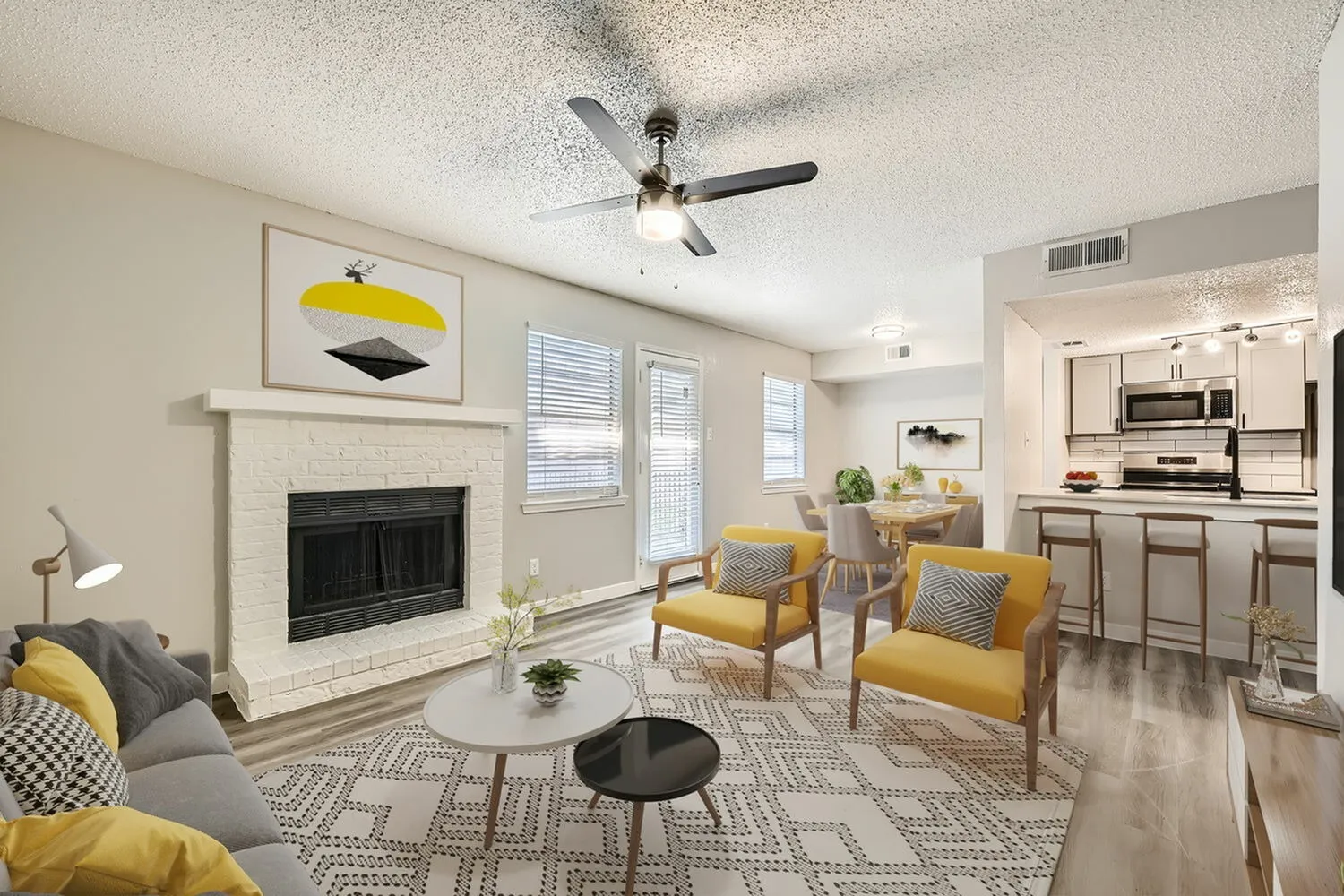 Open concept living area with gray sofa, yellow accent chairs, fireplace, ceiling fan, wood-style plank flooring, kitchen with stainless appliances visible