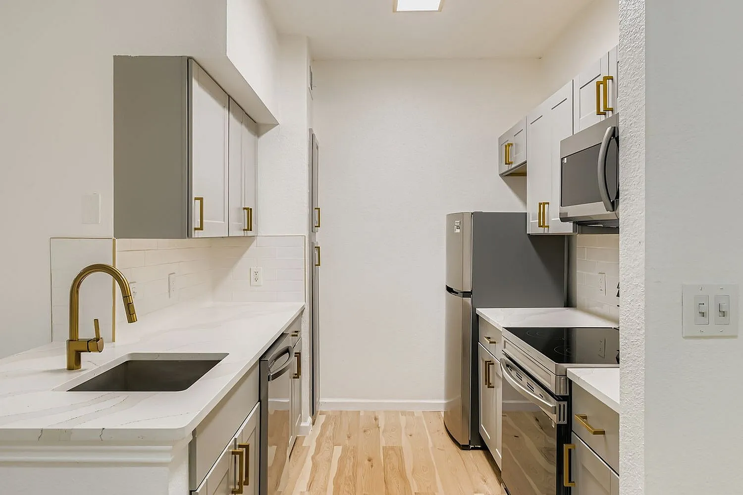 Galley kitchen, white shaker cabinetry, quartz countertops, brass fixtures, stainless appliances, wood-style plank flooring