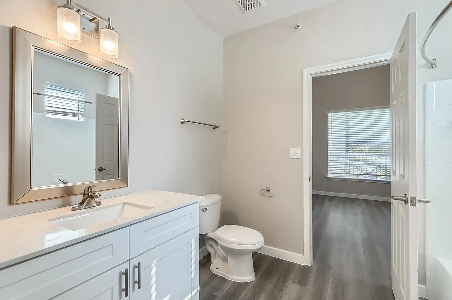 Primary bathroom, white vanity with quartz countertop, framed mirror, wood-style plank flooring, toilet, door to bedroom