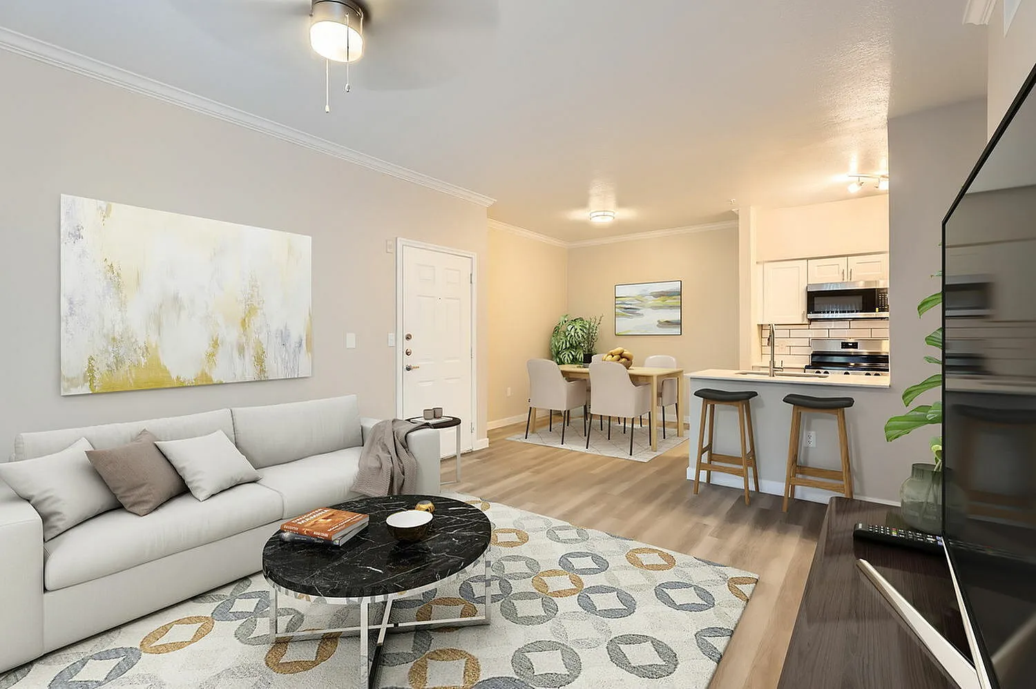 Living room with light gray sofa, abstract artwork, wood-style plank flooring, black round coffee table, patterned area rug, open kitchen with white cabinetry and bar seating