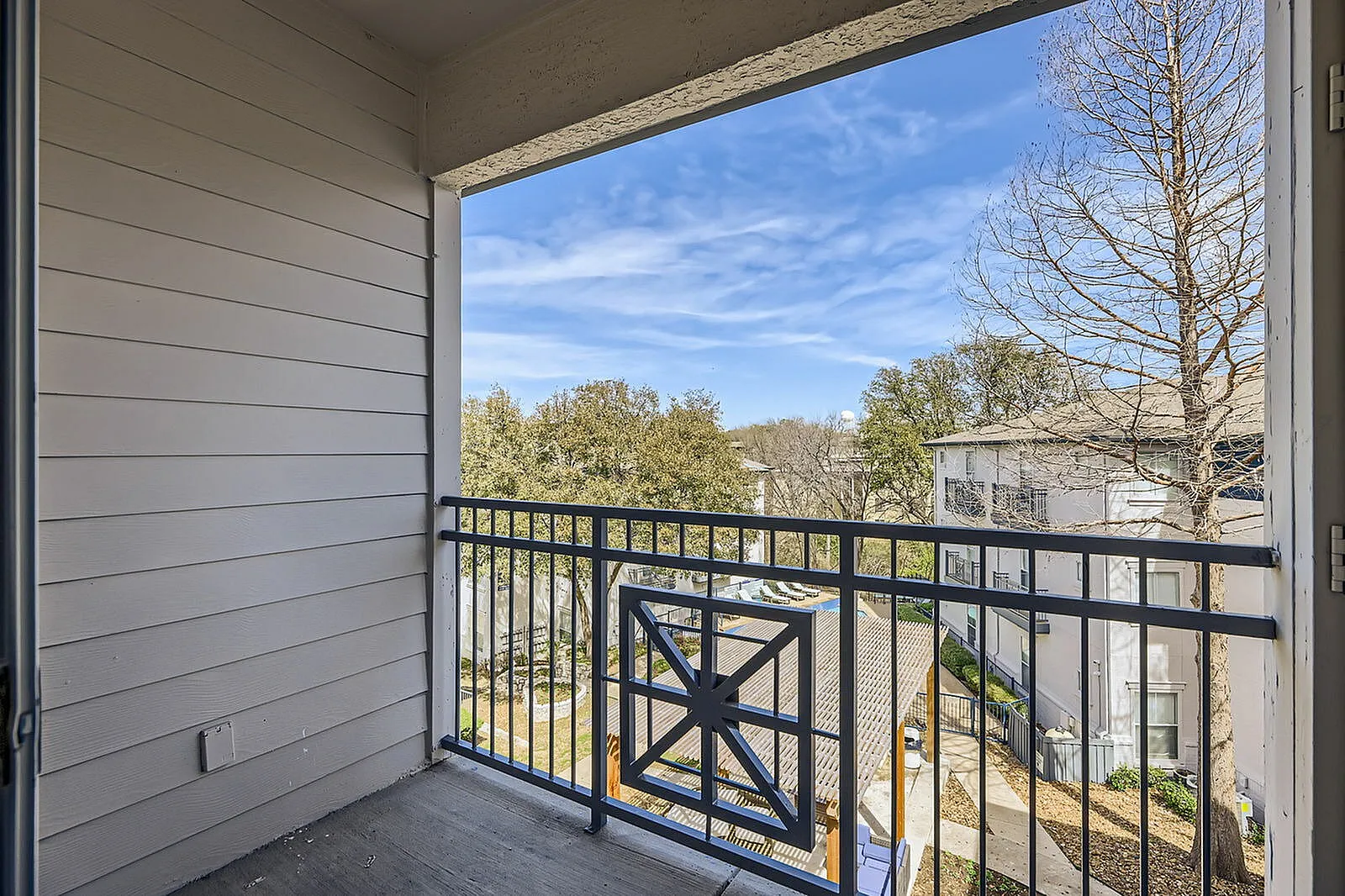 Balcony with black railing and gate, wood-style plank flooring, concrete slab, trees and sky visible