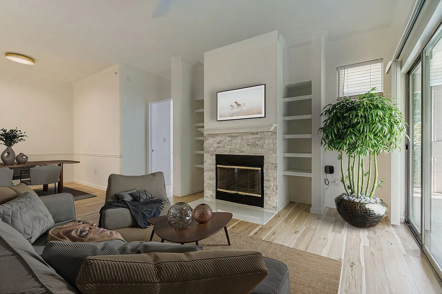 Living room, gray sectional sofa, wood-style plank flooring, stone fireplace with built-in shelving, potted plant