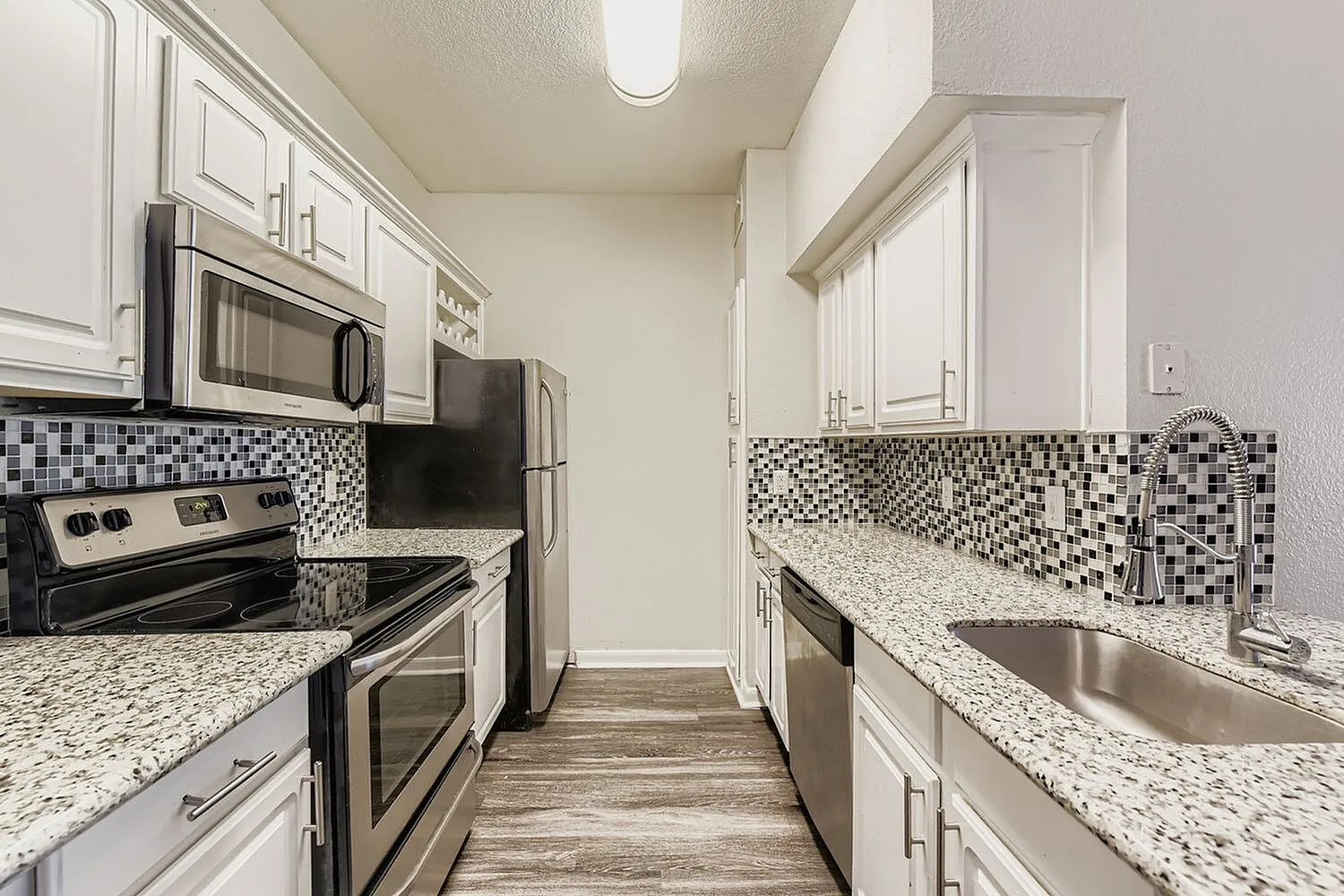 Galley kitchen, white shaker cabinetry, quartz countertops, stainless steel appliances, black and white mosaic tile backsplash, wood-style plank flooring