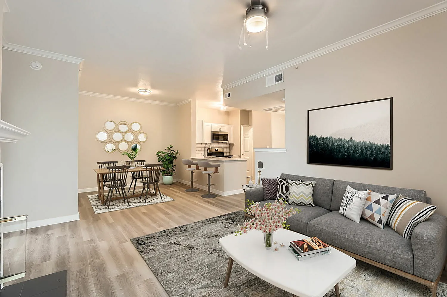 Open concept living and dining space, wood-style plank flooring, gray sofa with patterned pillows, white coffee table, dining chairs with round wall art