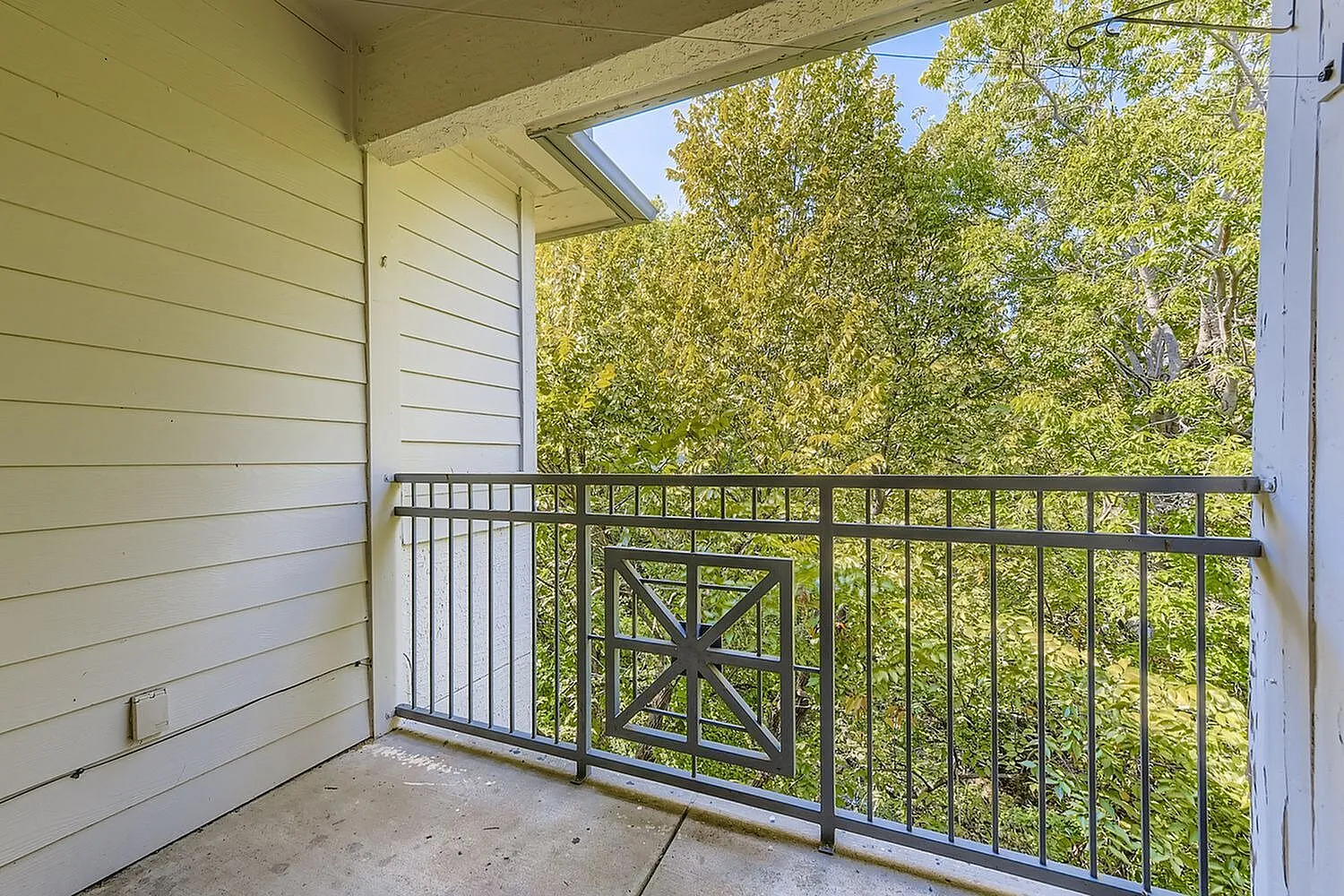 Balcony, metal railing with X-pattern gate, green tree canopy view, white siding