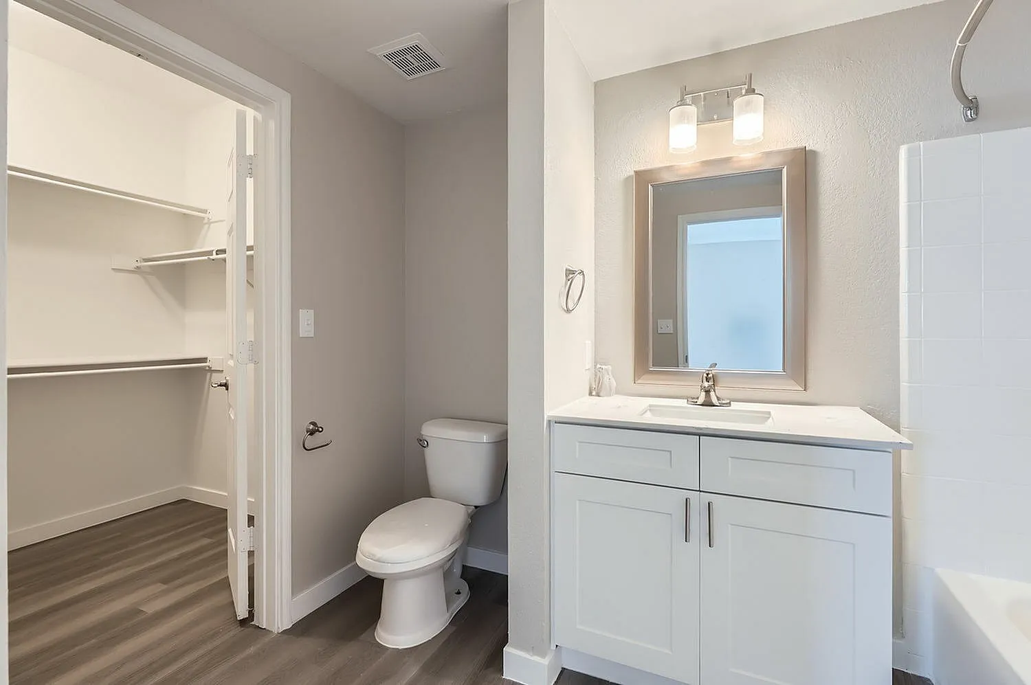 Primary bath, white cabinetry, quartz countertop, framed mirror, wood-style plank flooring, walk-in closet