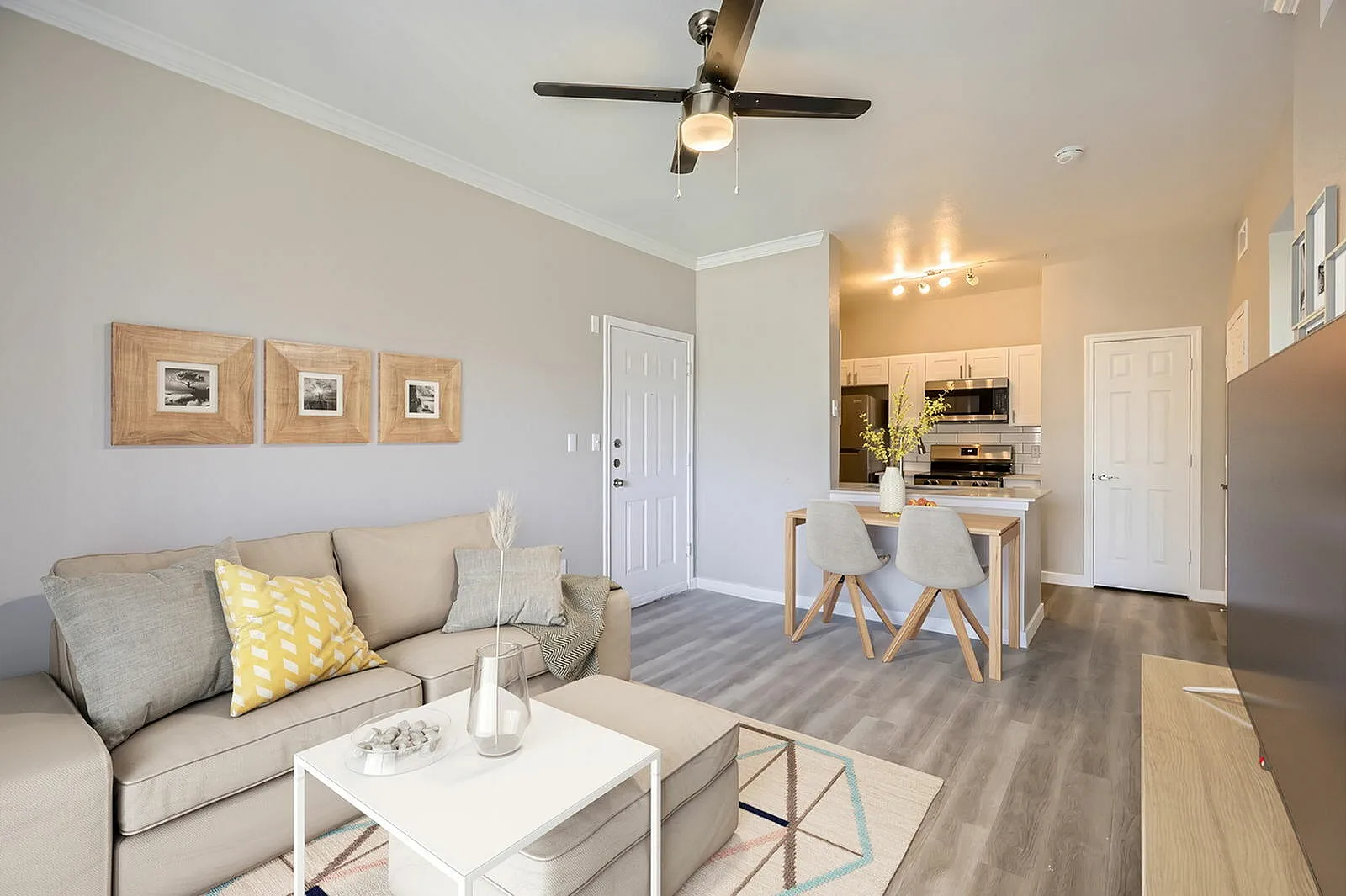 Living room with wood-style plank flooring, gray sofa, ceiling fan, kitchen with white cabinetry
