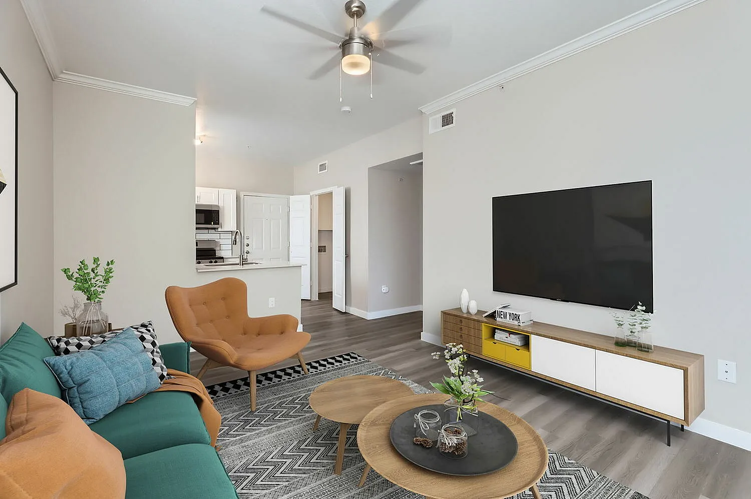 Living room with wood-style plank flooring, modern tan leather chairs, teal sectional, ceiling fan, TV on media console, open kitchen with white cabinetry