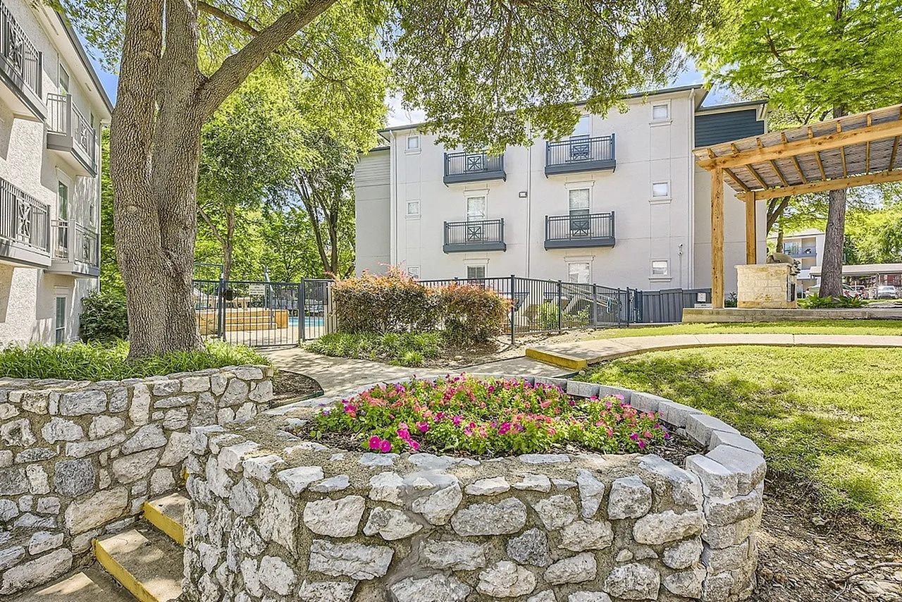 Courtyard with stone planter beds, flowering plants, mature trees, building facade, wooden pergola