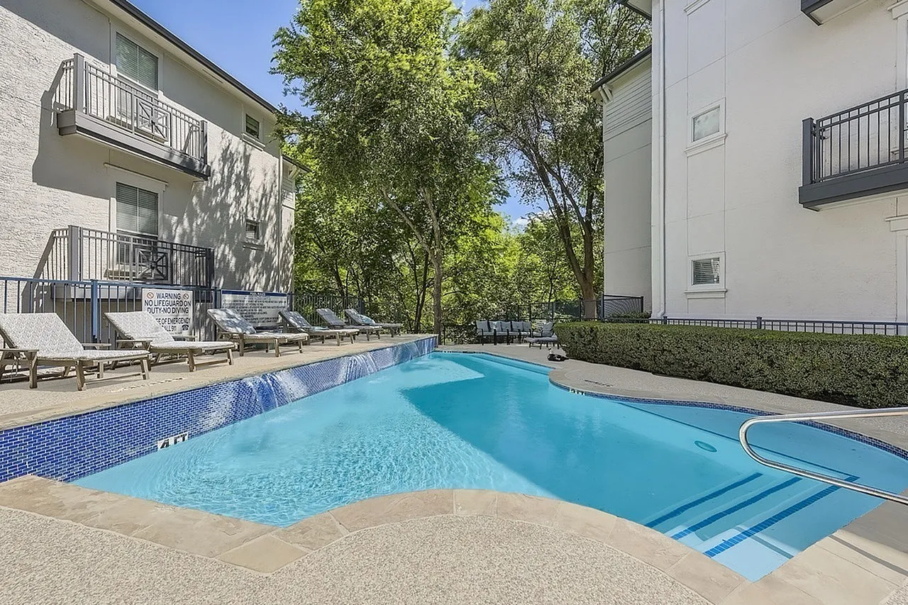 Swimming pool with bright turquoise water, concrete deck, residential buildings, shade trees, lounge seating