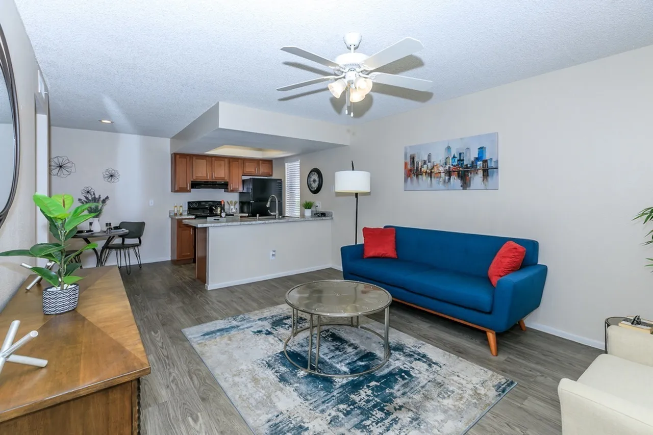 Open-concept living room, blue sofa with red pillows, wood-style plank flooring, kitchen with wood cabinetry, ceiling fan