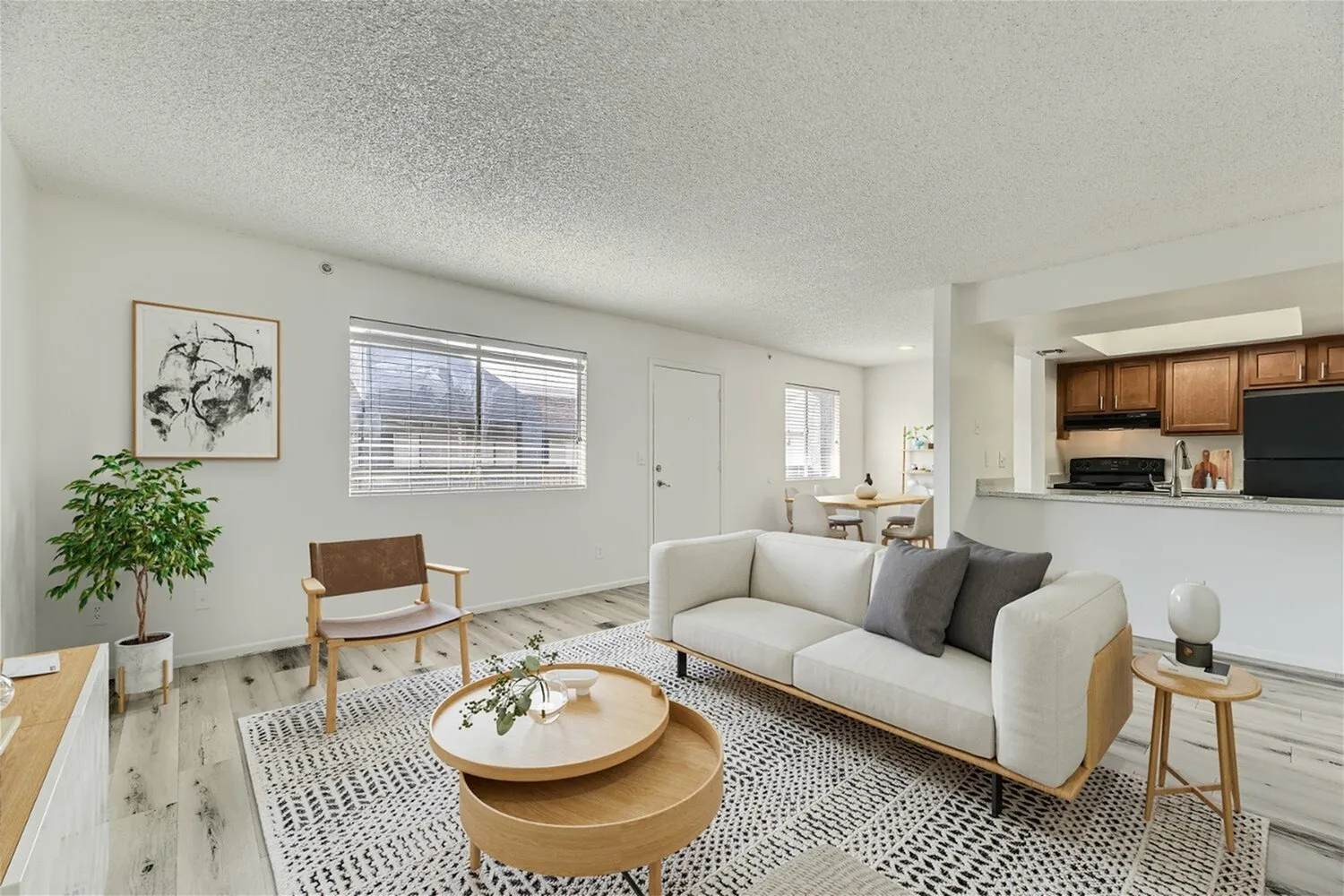 Living room with cream sofa, wood-style plank flooring, geometric area rug, round wood coffee table, open kitchen with wood cabinetry and black appliances