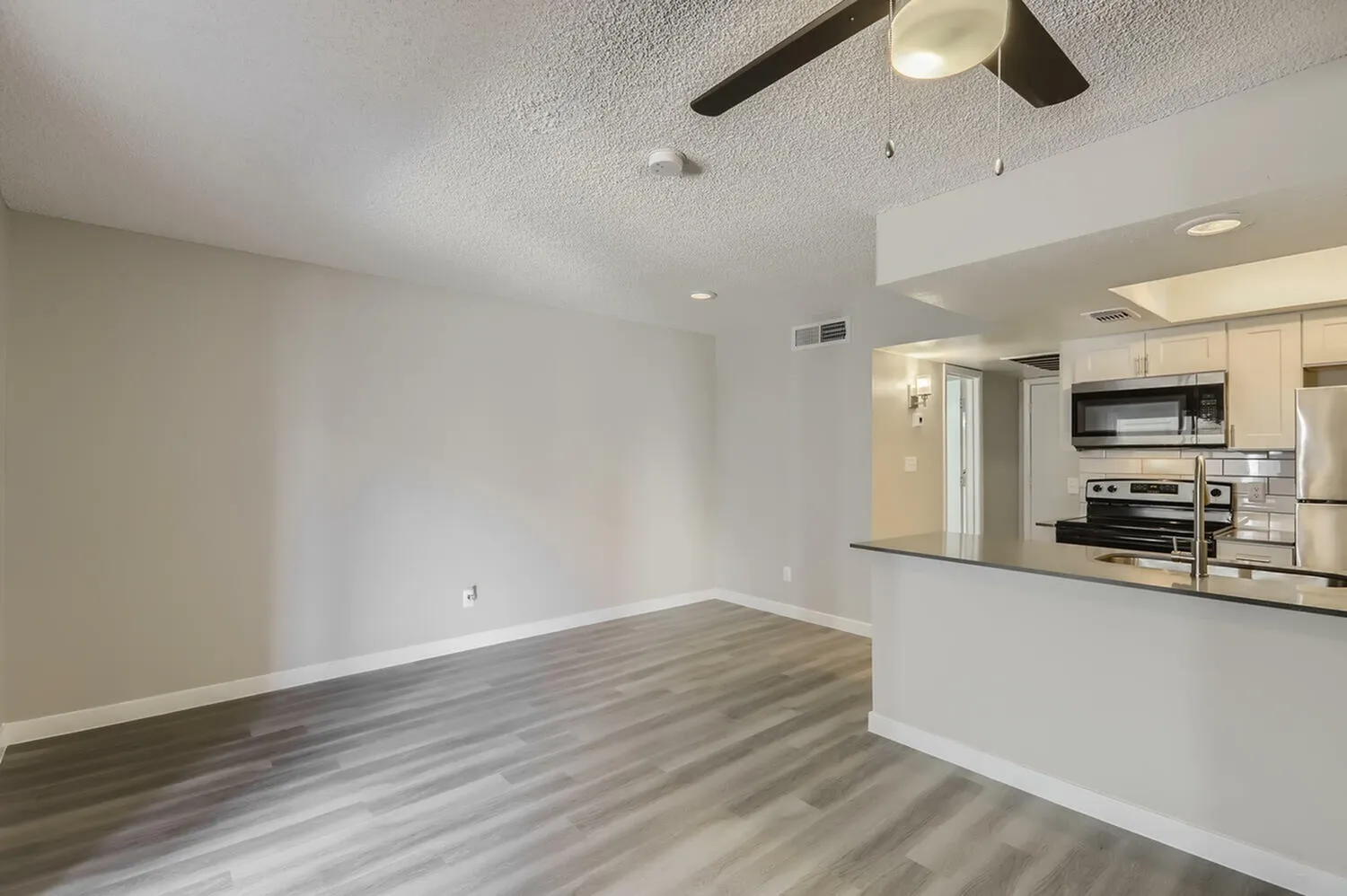 Living room with wood-style plank flooring, ceiling fan, open kitchen with stainless appliances and white cabinetry