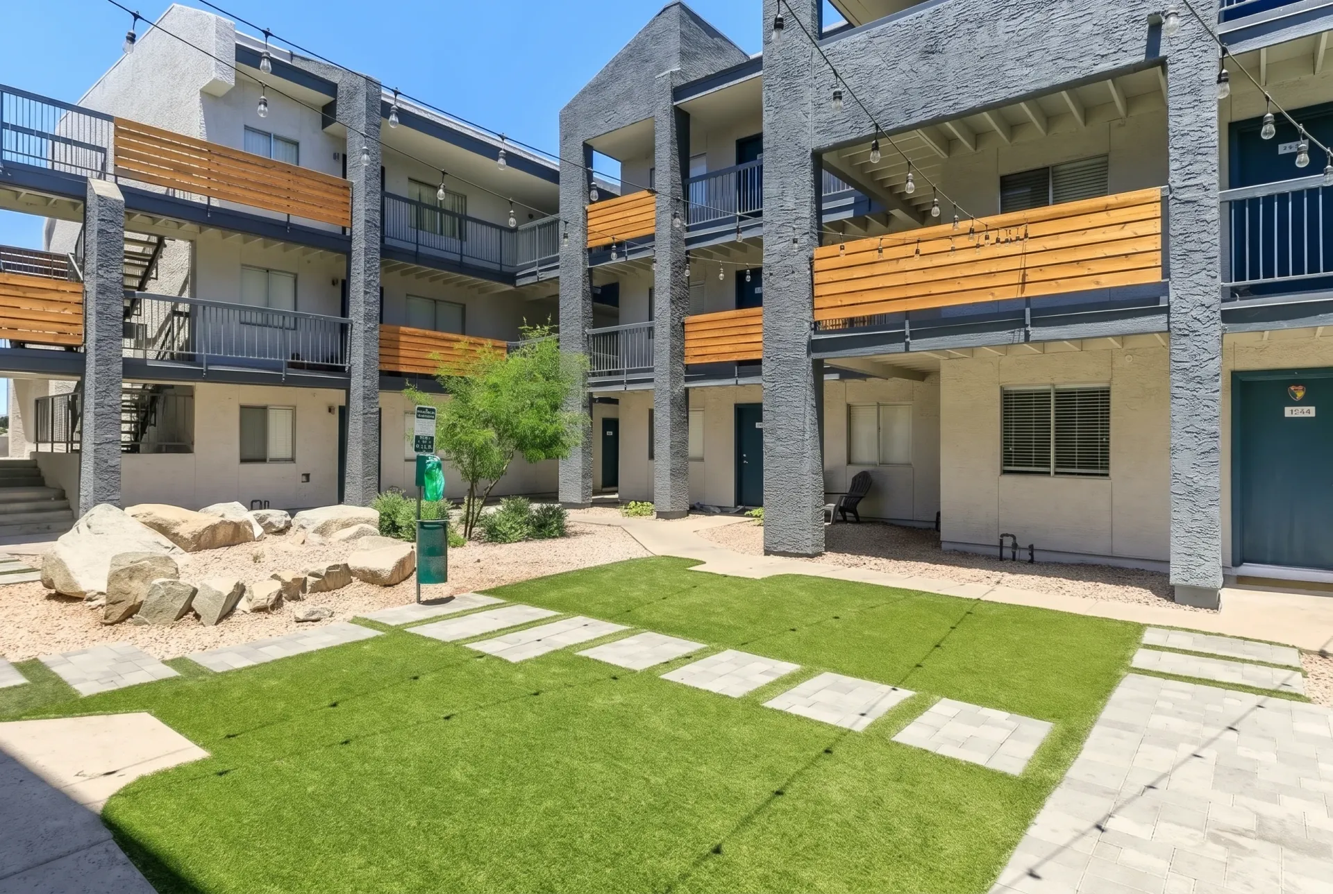 Courtyard with green lawn, stepping stone pathway, decorative boulders, concrete planter, multi-story residential buildings with wood siding and stone exterior accents