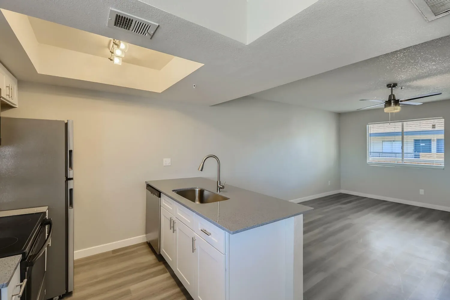 Kitchen with white cabinetry, stainless steel sink, wood-style plank flooring, living room with ceiling fan and windows
