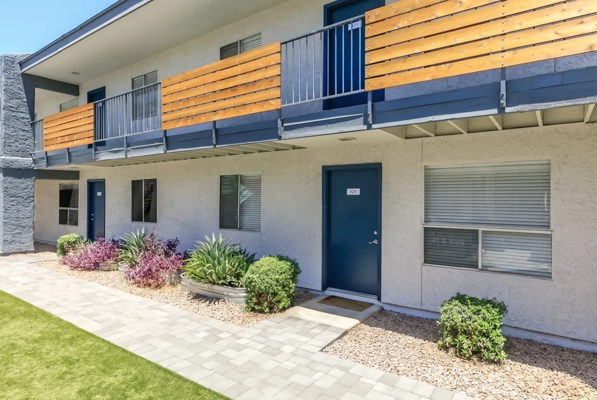 Ground level patio with stucco walls, dark blue entry door, shrub landscaping, wood-style siding balcony above