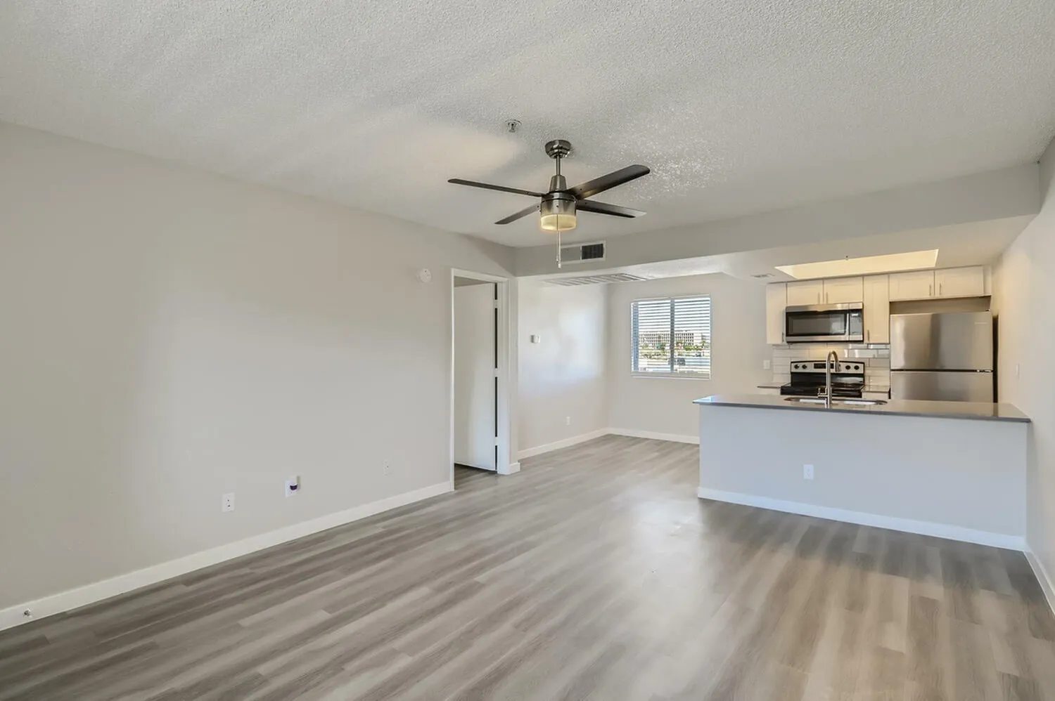 Living room with wood-style plank flooring, ceiling fan, kitchen with white cabinetry, stainless steel appliances