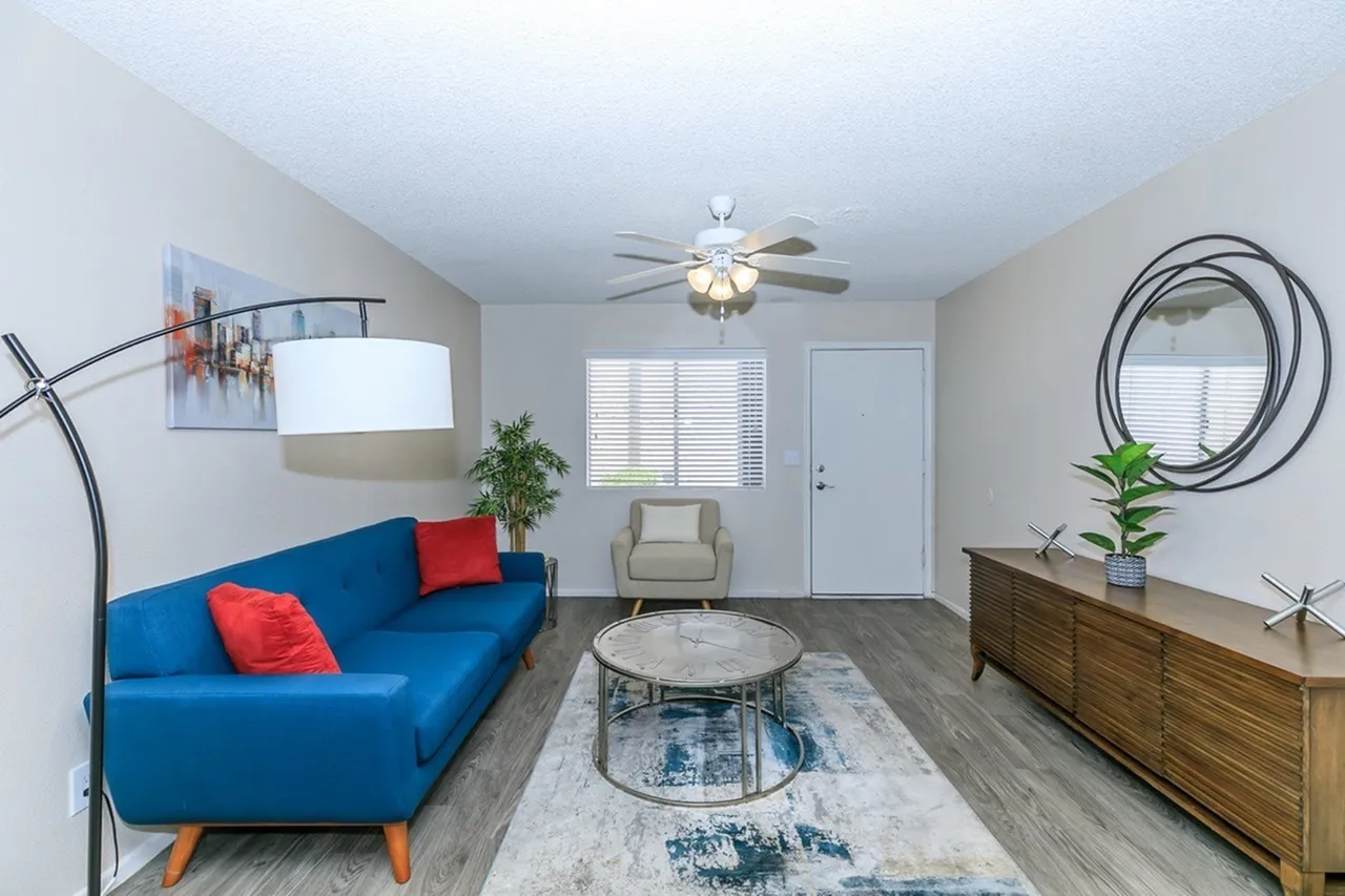 Living room, blue sofa with red pillows, wood-style plank flooring, ceiling fan, round mirror accent