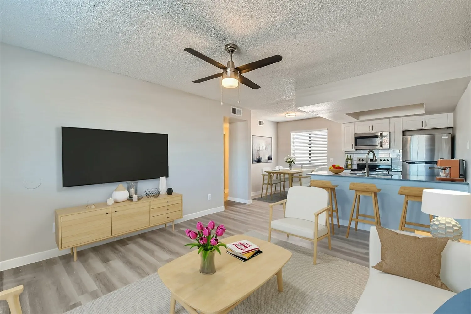 Open concept living room, wood-style plank flooring, ceiling fan, mounted flat-screen TV, wood furniture, kitchen with stainless appliances visible
