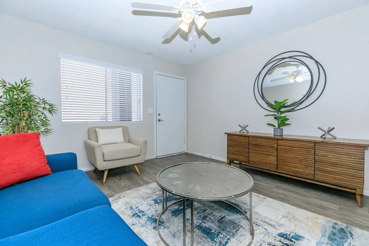Living room, blue sofa with red pillow, wood-style plank flooring, mid-century chair, round glass coffee table, wood dresser, circular wall mirror, ceiling fan, white shuttered window