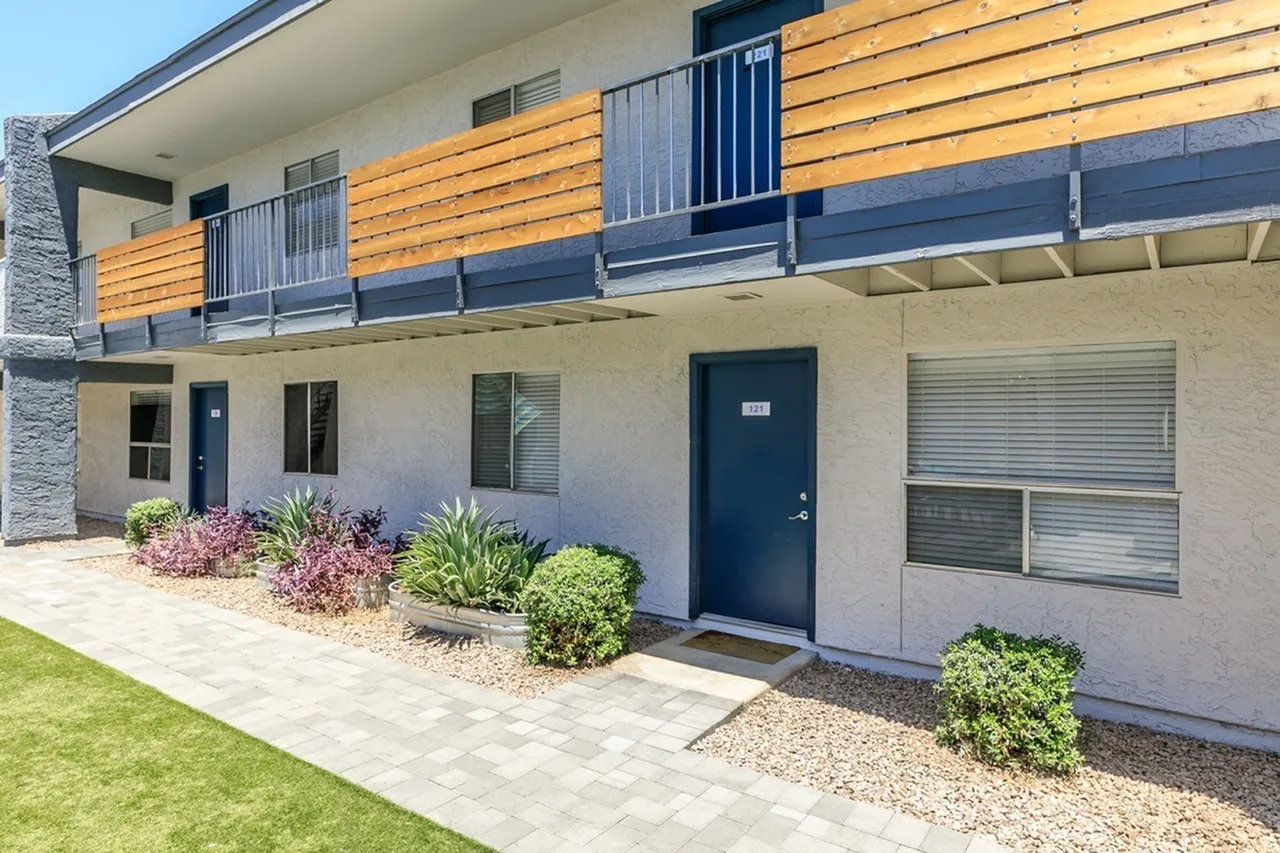 Ground level patio with stucco walls, dark blue entry door, shrub landscaping, wood-style siding balcony above