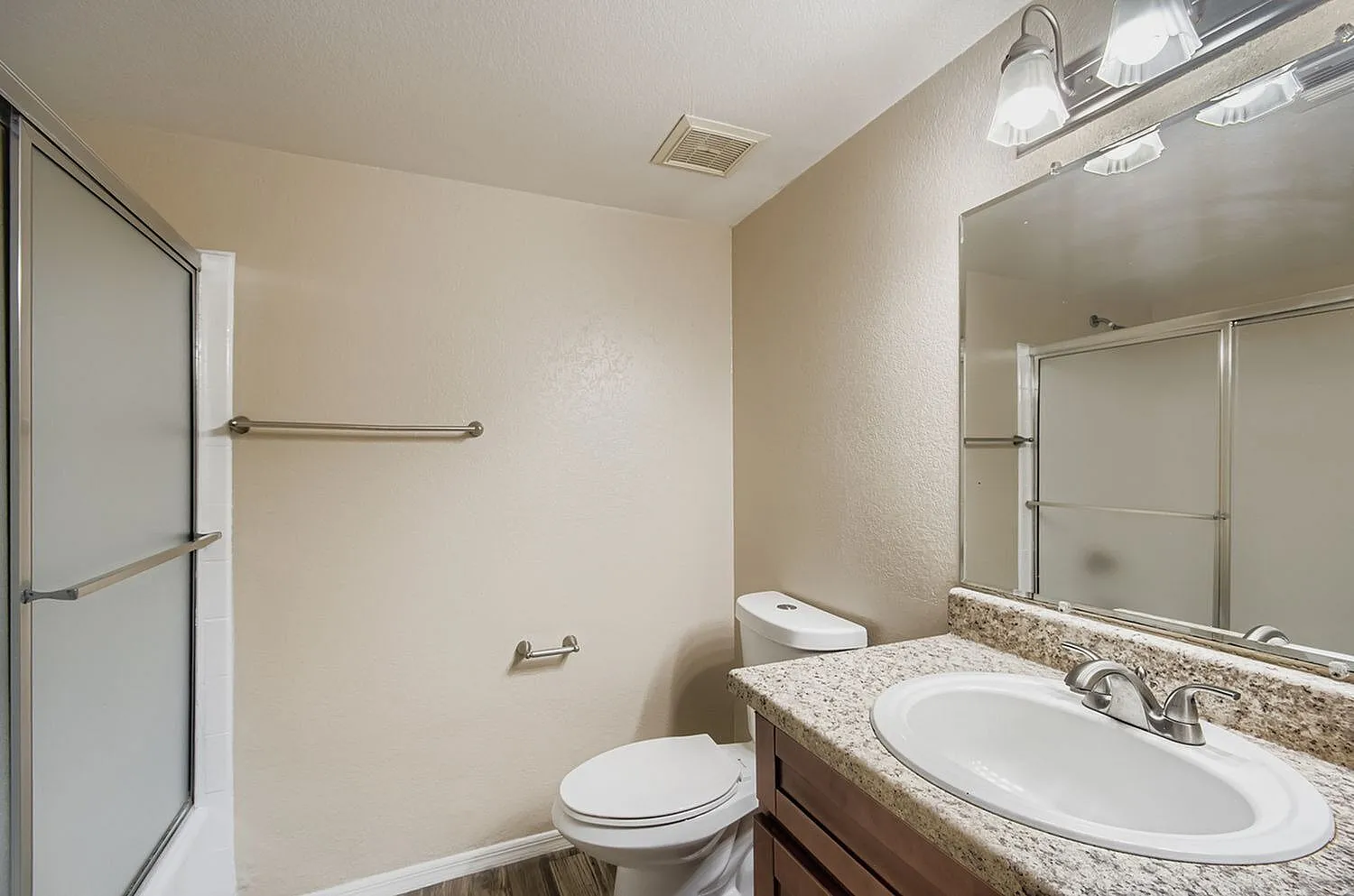 Bathroom with granite countertop, white oval sink, large mirror, toilet, shower with glass enclosure, wood-style plank flooring