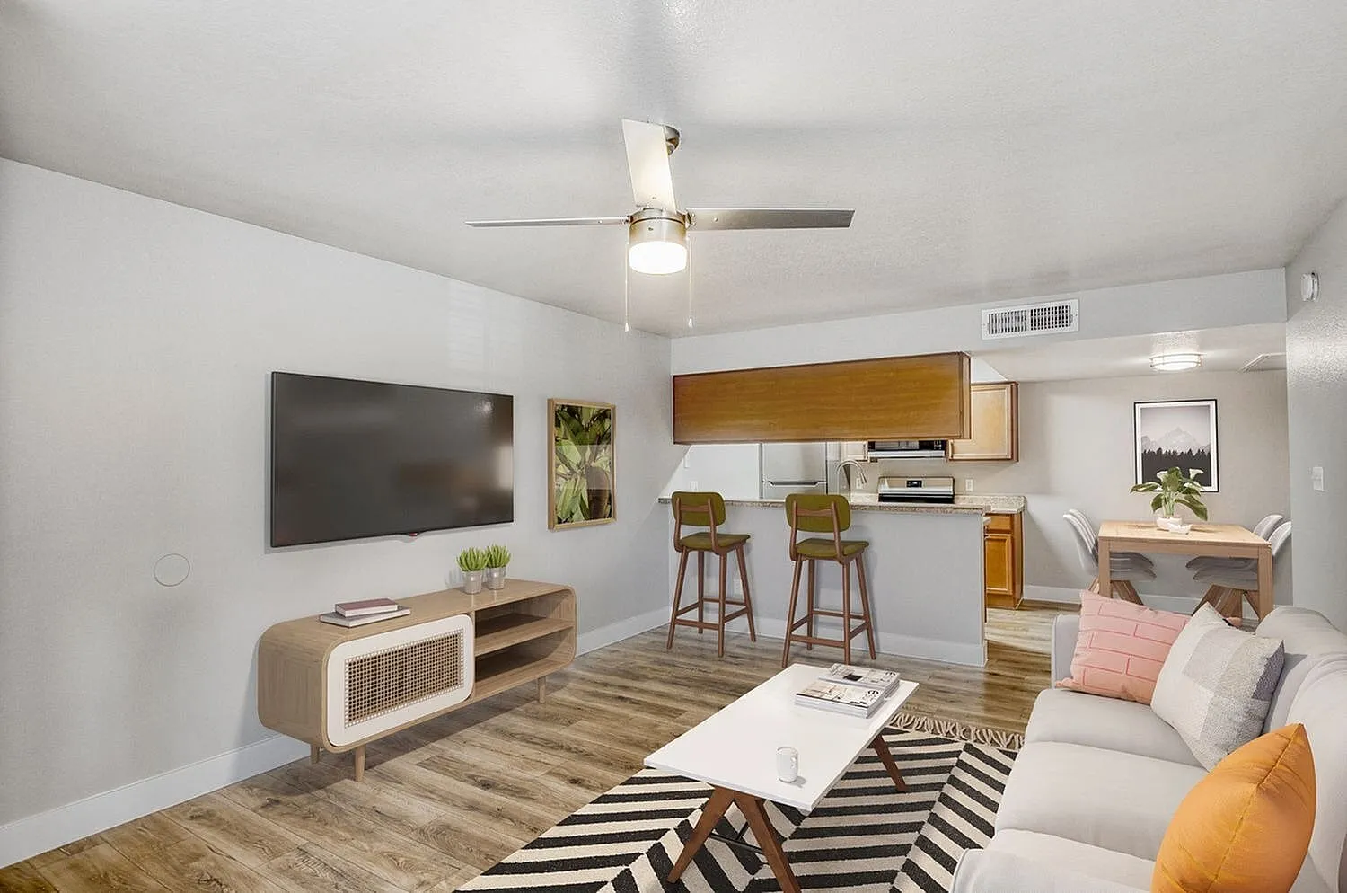 Living room with wood-style plank flooring, ceiling fan, mounted TV, kitchen bar seating, wood cabinetry