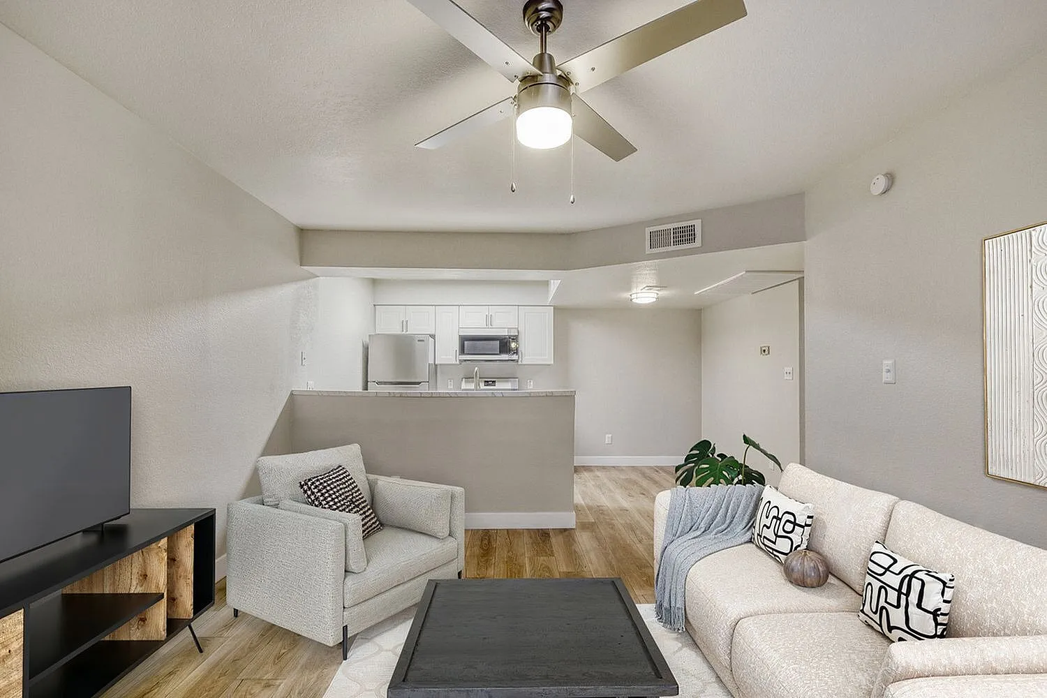Living room with beige sofa, gray armchair, wood-style plank flooring, ceiling fan, kitchen with white cabinetry
