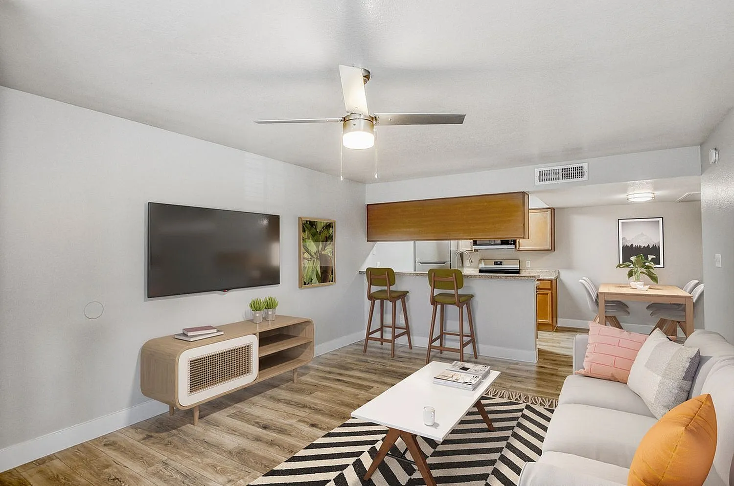 Living room with wood-style plank flooring, ceiling fan, flat-screen TV, contemporary media console, kitchen with wood cabinetry, bar seating