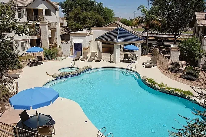 Resort-style pool with curved shape, blue umbrellas, cabana with dark roof, spa, loungers
