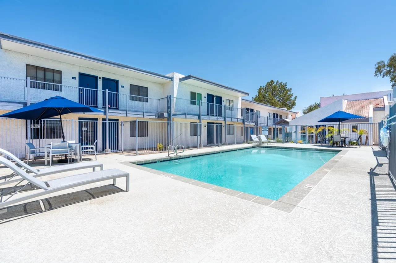 Resort-style pool area, lounge chairs, blue umbrellas, multi-unit building, clear sky