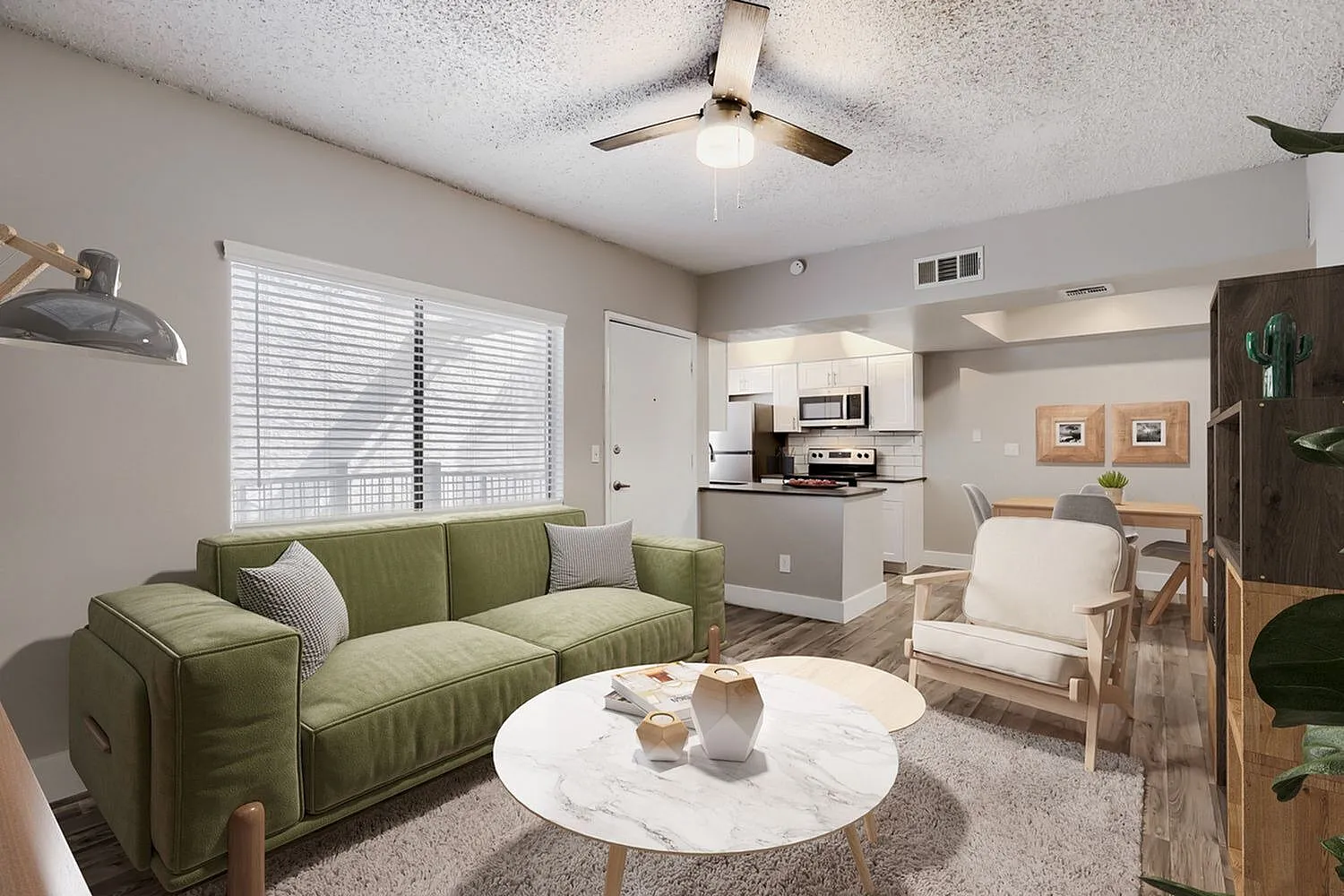Living room with olive green sofa, white round marble table, ceiling fan, wood-style plank flooring, open kitchen with white cabinetry