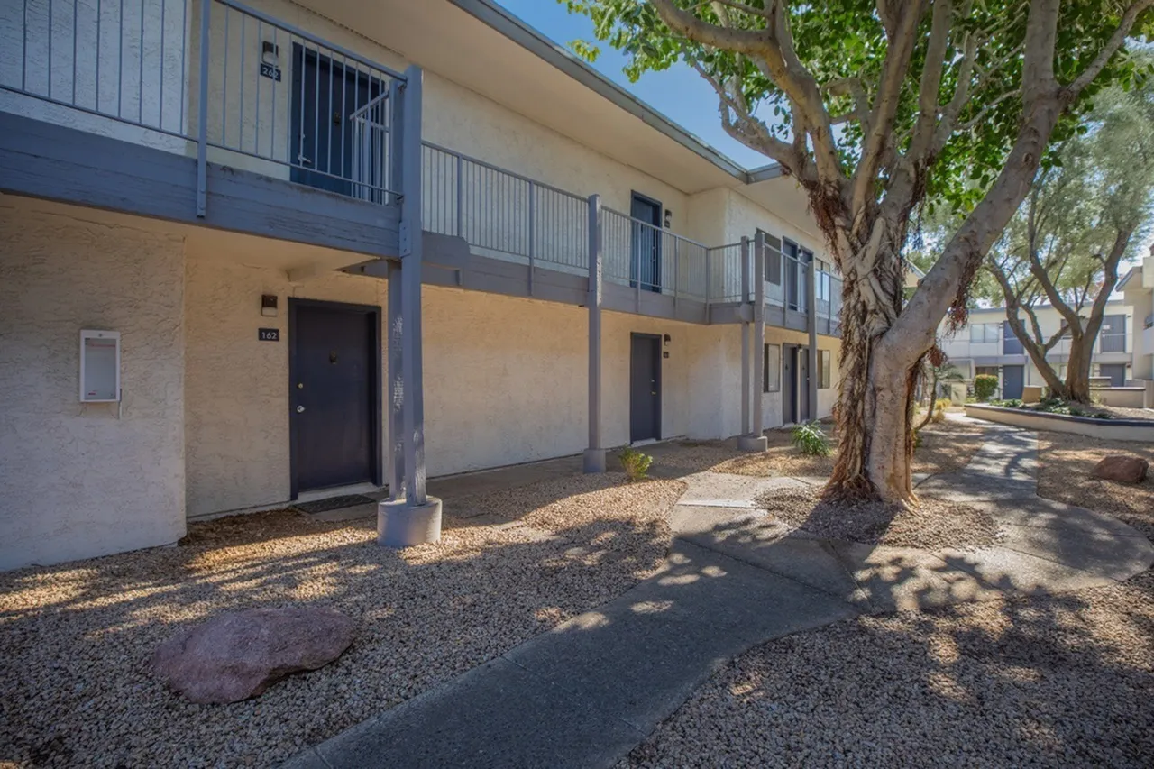 Exterior courtyard, two-story residential building, blue metal railings, mature tree, gravel ground