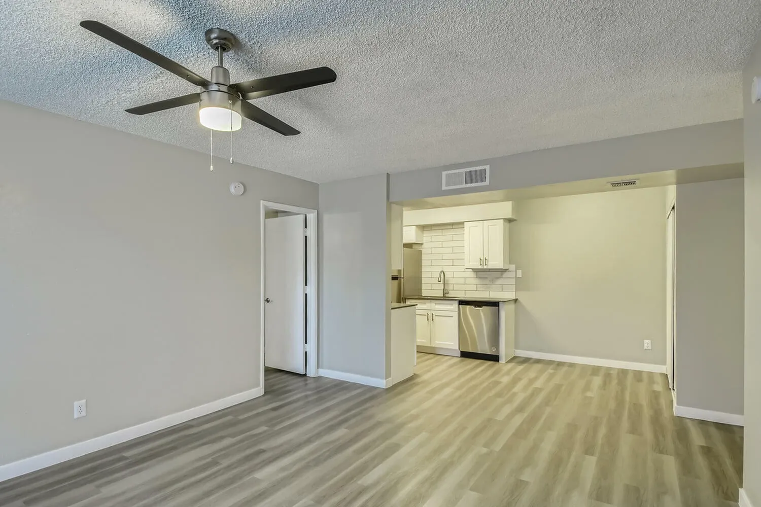 Living area, wood-style plank flooring, ceiling fan, open kitchen with white cabinetry