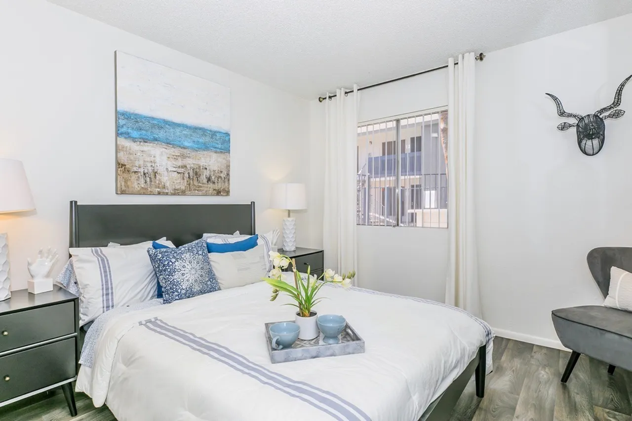 Primary bedroom, dark headboard, blue accents, wood-style plank flooring, window with sheer curtains