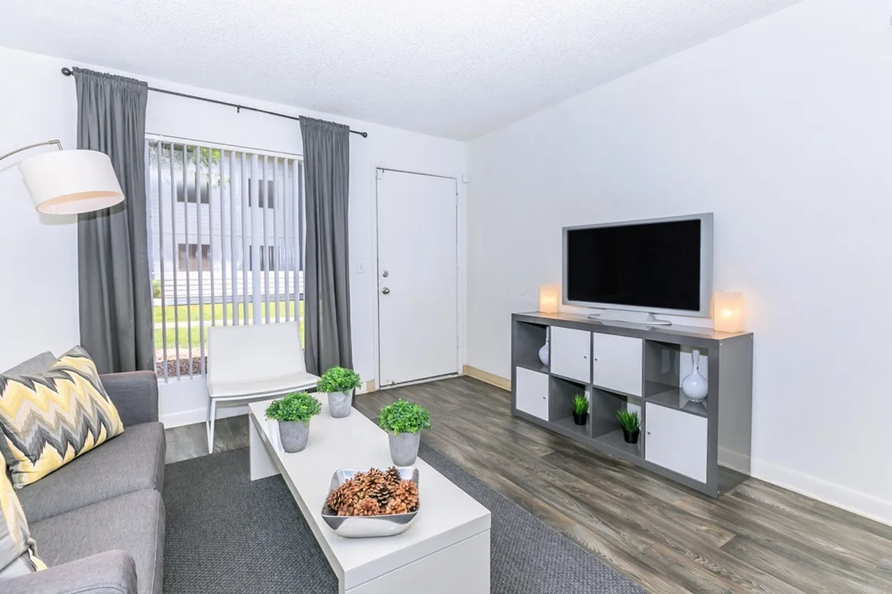 Living room, wood-style plank flooring, gray upholstered sofa, flat-screen TV mounted on media unit with storage cubbies, white coffee table, potted green plants, gray curtains, floor lamp