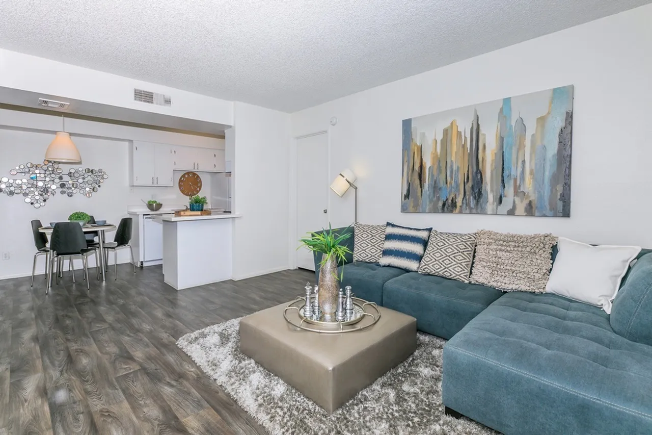Living room with teal sectional sofa, wood-style plank flooring, open kitchen with white cabinetry, city skyline artwork