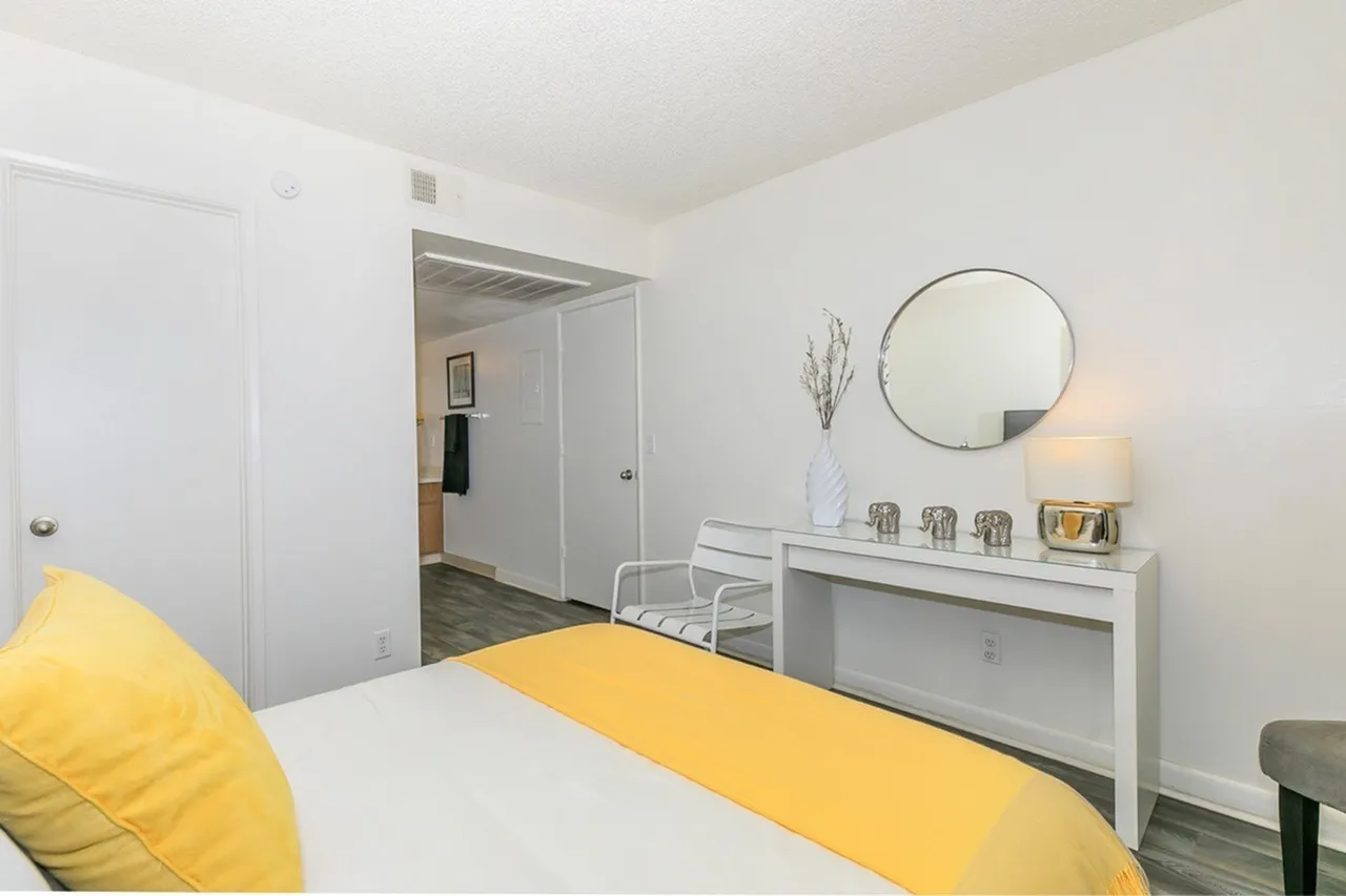 Bedroom, wood-style plank flooring, white bedding, yellow throw pillows, white vanity with round mirror