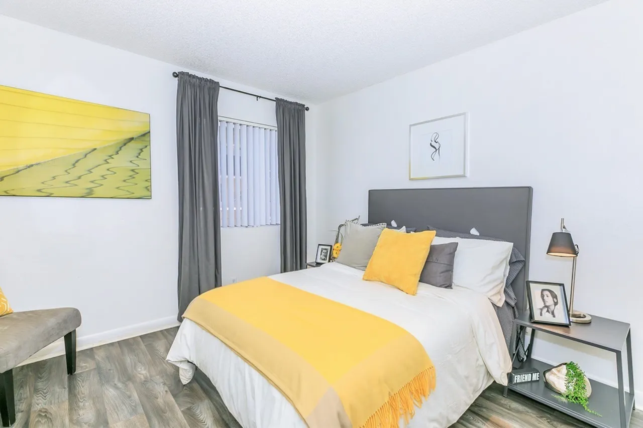 Primary bedroom, gray upholstered headboard, yellow accent bedding, gray curtains, wood-style plank flooring