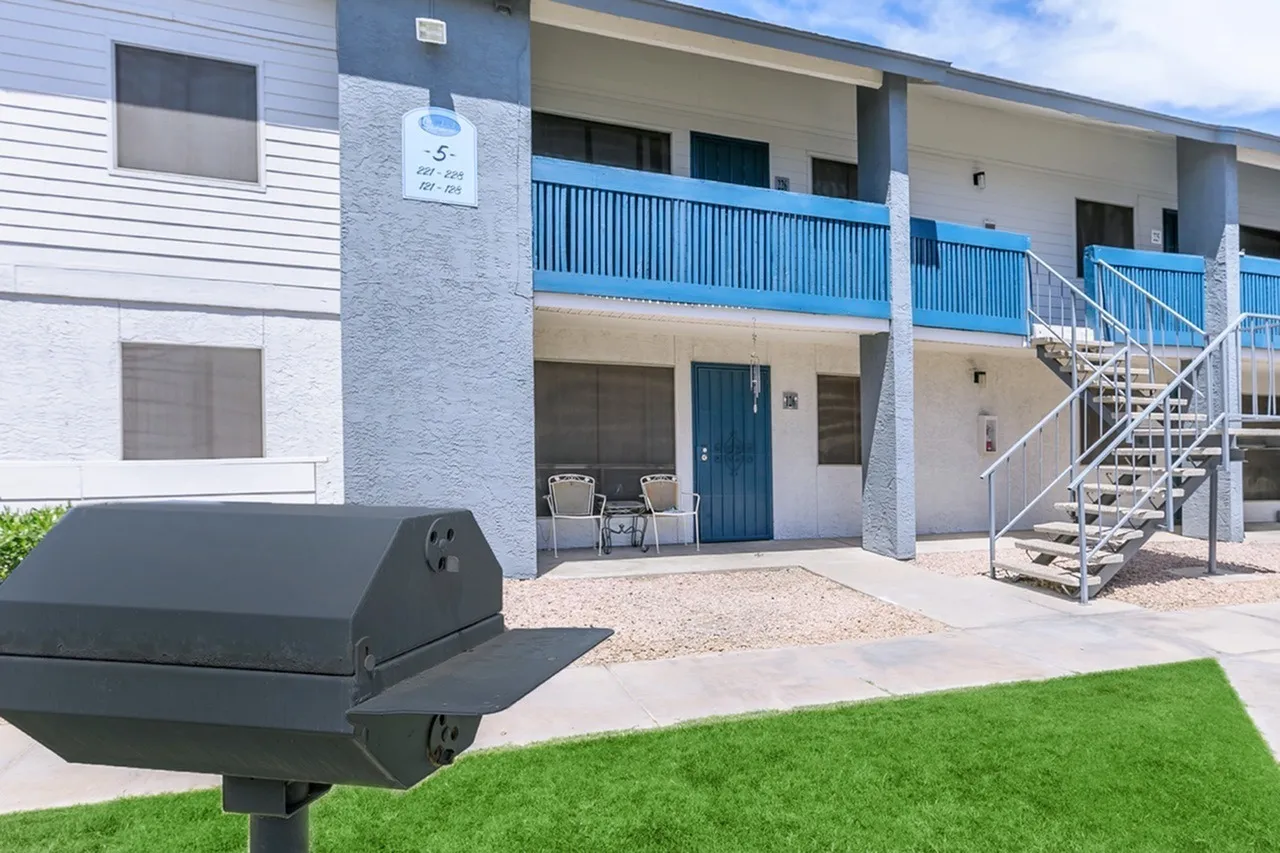 Multi-story apartment building, blue metal railings, white and tan exterior, metal staircase, gravel courtyard, green lawn