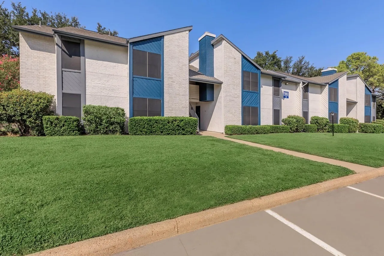 Modern apartment building with beige brick facade, blue glass windows and accents, manicured lawn, landscaping