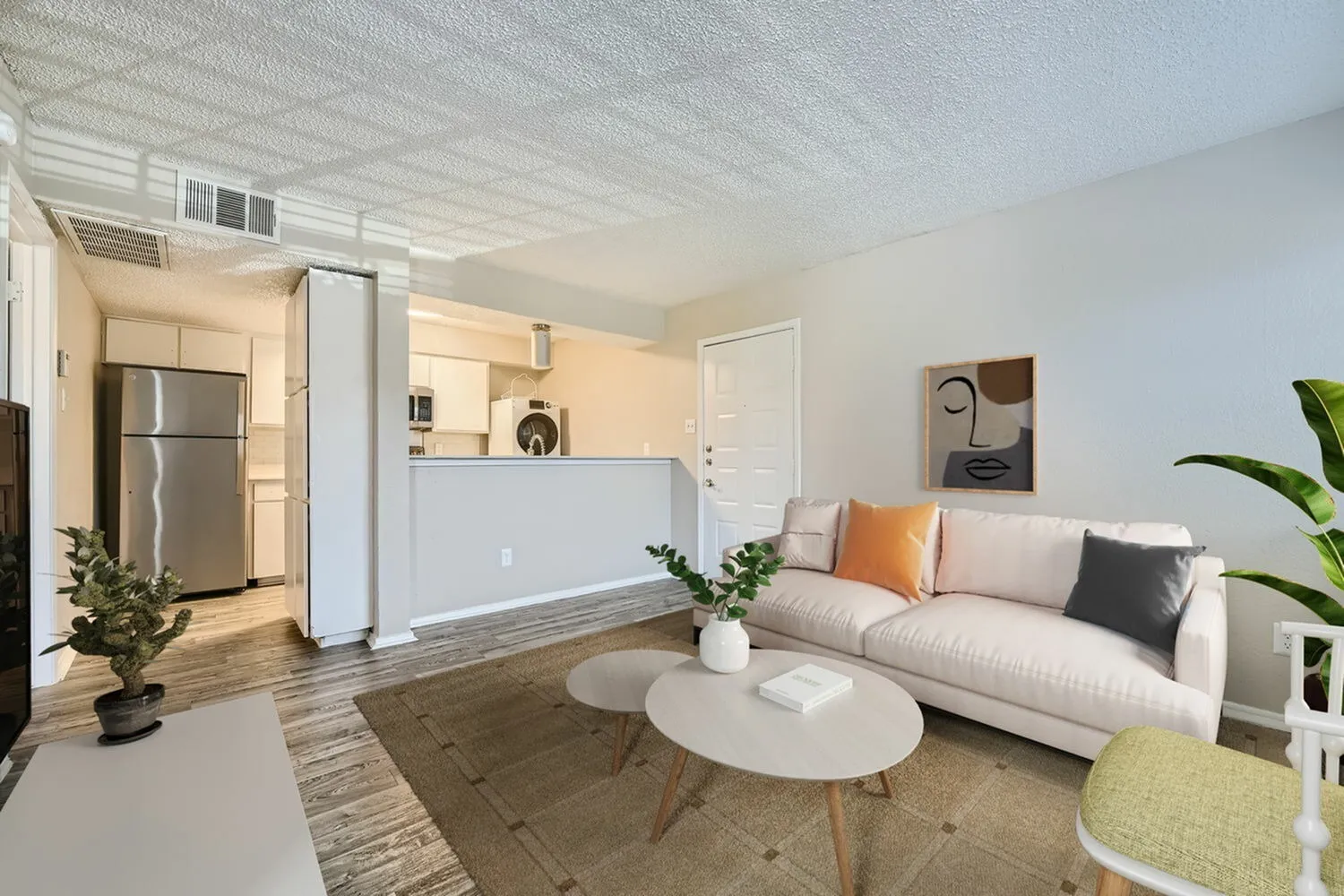 Open concept living and kitchen area, wood-style plank flooring, stainless appliances, cream sofa with colorful pillows, white round coffee table