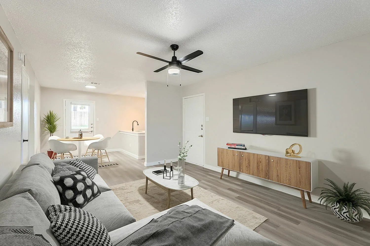 Open living room with gray sofa, wood-style plank flooring, ceiling fan, flat-screen TV mounted on white wall, wood media console, white kitchen with island in background