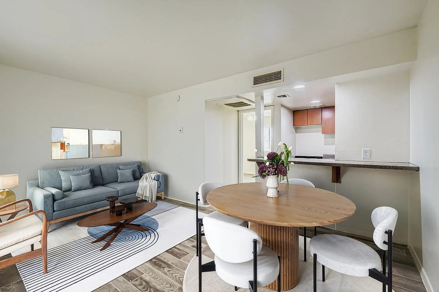 Open concept living room with blue sofa, wood-style plank flooring, oval wood dining table, white upholstered chairs, kitchen with stainless appliances visible in background