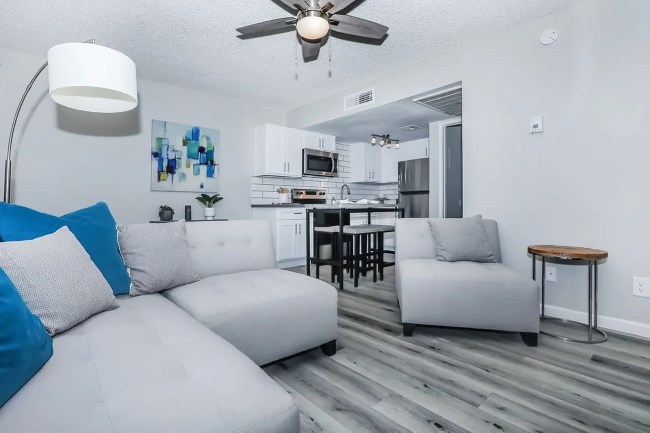 Living room with light gray sectional sofa, wood-style plank flooring, ceiling fan, arc floor lamp, dining area with black table and stools, kitchen with white shaker cabinetry and stainless appliances