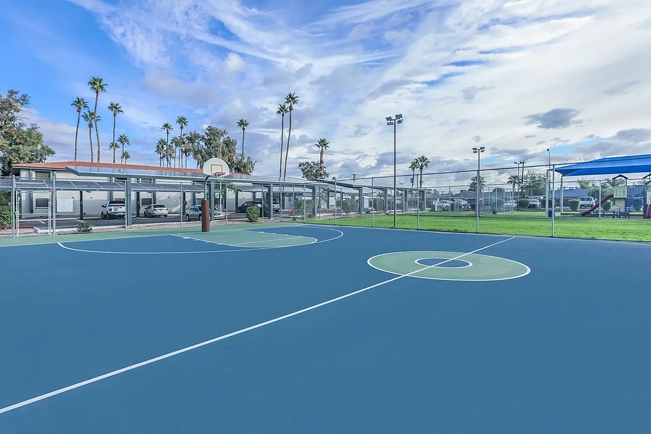 Basketball court with blue playing surface, center court markings, palm trees, outdoor lighting, built structures