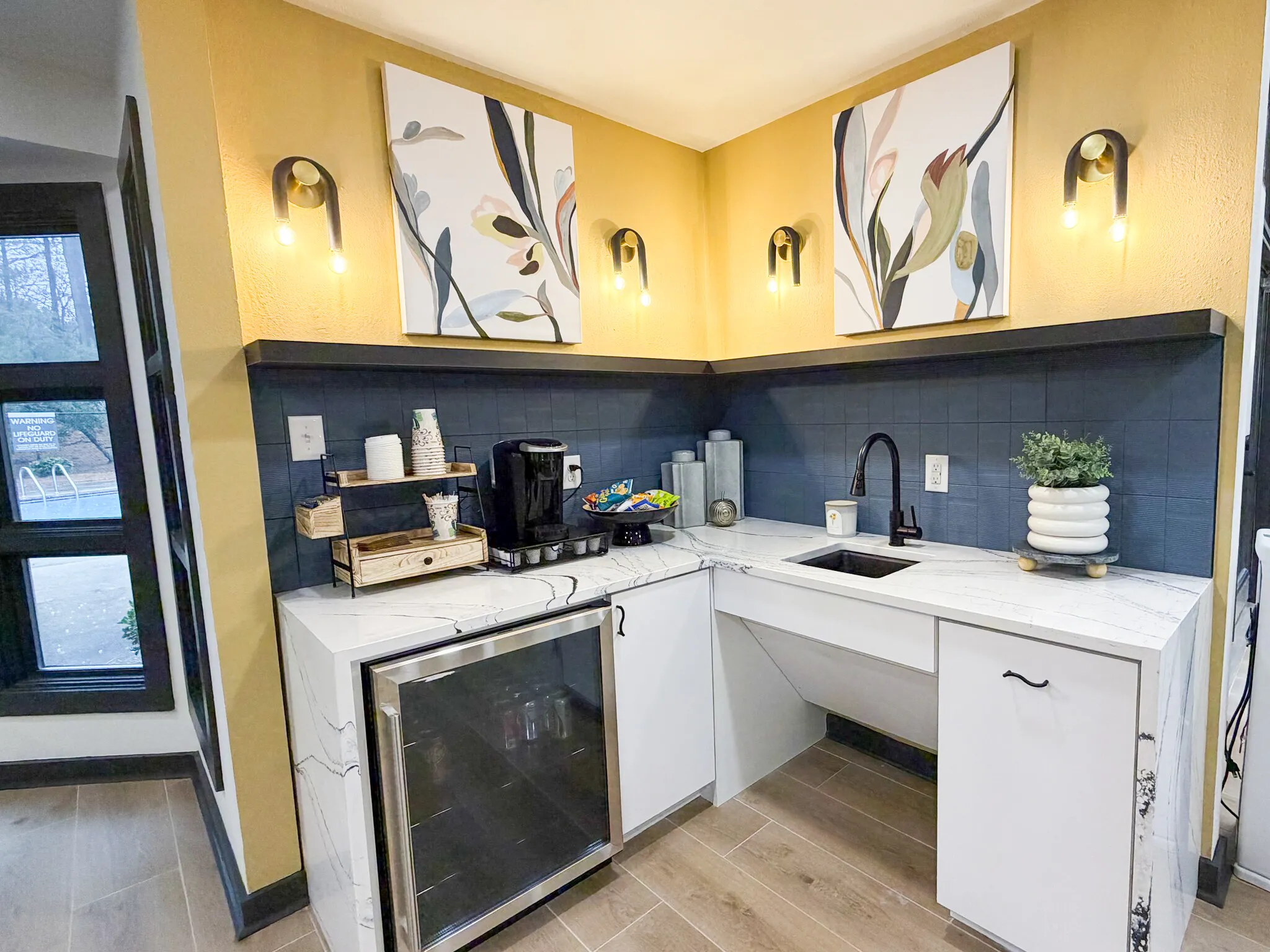 Break room with yellow walls, blue accent backsplash, white cabinetry, quartz countertops, sink, stainless appliances, wood-style plank flooring