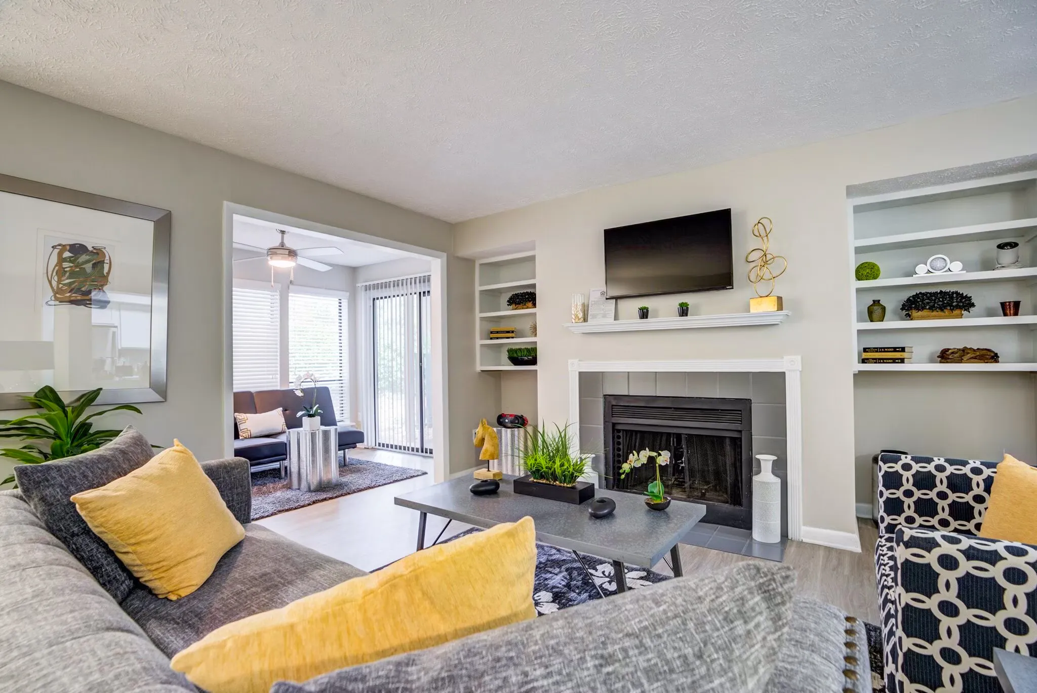 Living room, gray sofa with yellow pillows, fireplace with black surround, wall-mounted TV, shelving, glass doors to sunroom