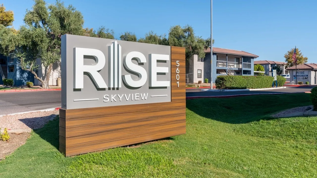 Monument sign with Rise Skyview logo, wood-style accent panel, green lawn, apartment buildings, trees